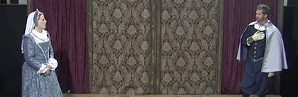A photo of Gallia and Cordella standing across from each other. Cordella’s hands are folded in front of her. Gallia has a hand on his heart.