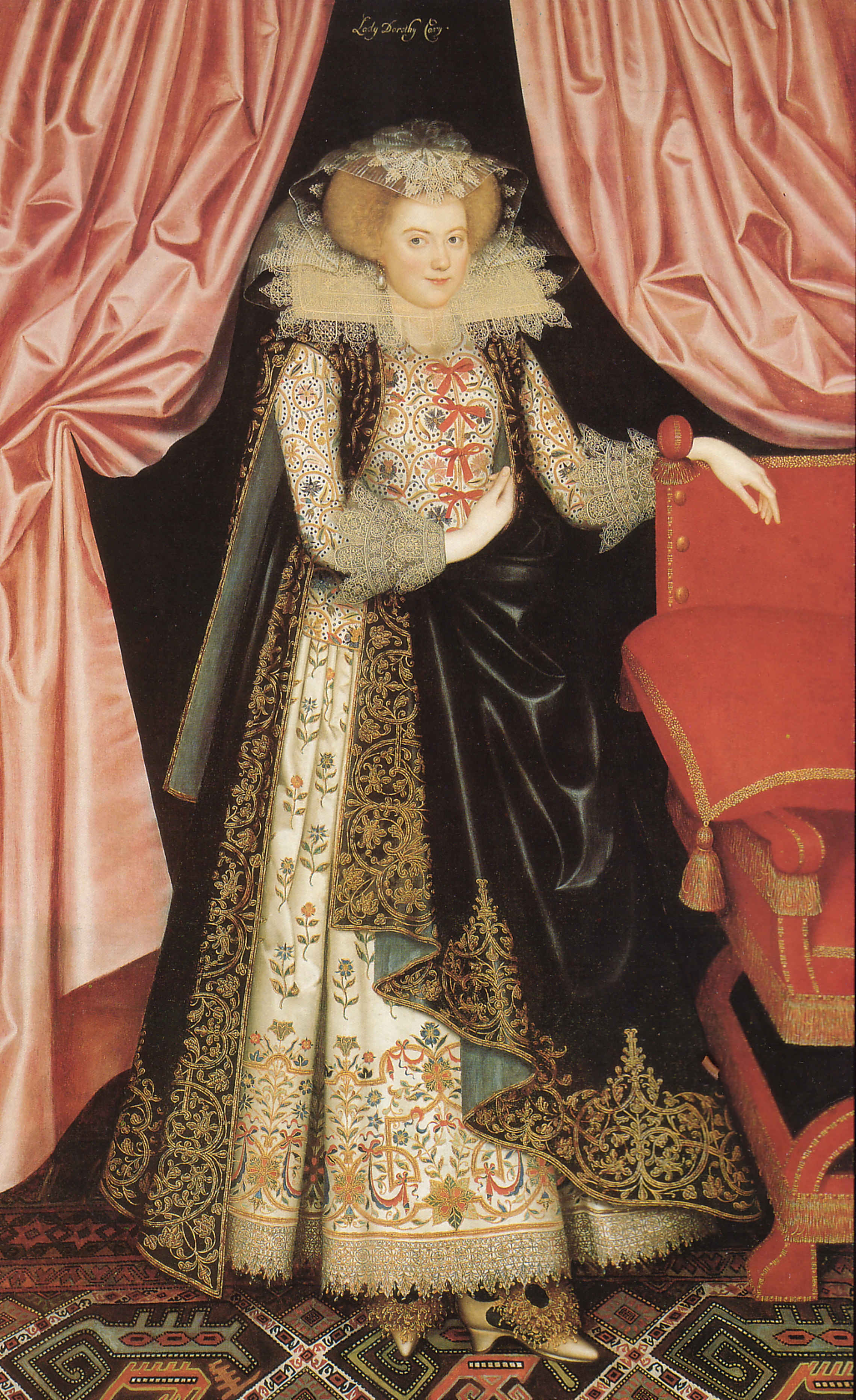 Image of an ornately dressed woman standing in between two pink curtains. Her hand rests on a pink chair, and her dress is made of a highly decorated cream underlayer and what looks to be a black velvet overlayer with gold embroidery.