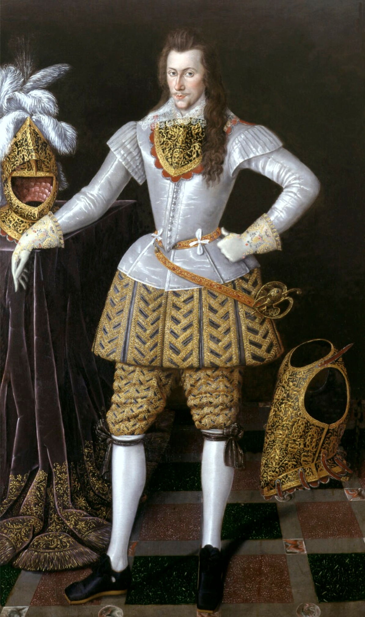 An oil on canvas painting of Henry Wriothesley. He stands with one white-gloved fist on his hip and the other arm resting on a black velvet draped table. He is dressed in a white shirt with a black, gold, and orange collar over his chest. His brown hair reaches the bottom of his chest. His legs are covered in a black and gold skirt, similar pants, and white socks that are tied with garters at the knee. A sword hangs at his hip, tied by a belt. Beside him on the left table is an ornate gold helmet with white feathers pluming from the top. On his right, a gold chestplate sits on the floor.