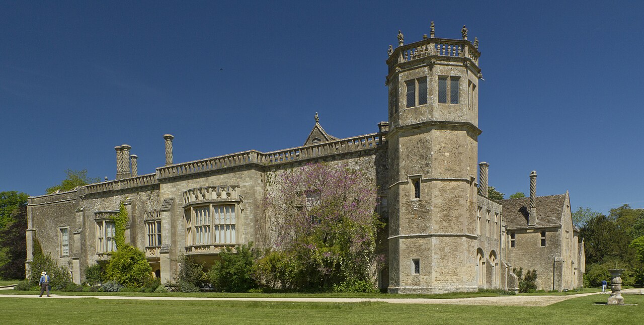 A photograph of a large, sandy colored building with a turret on the corner facing the photographer and large bay windows. Plants and ivy creep up its walls.
