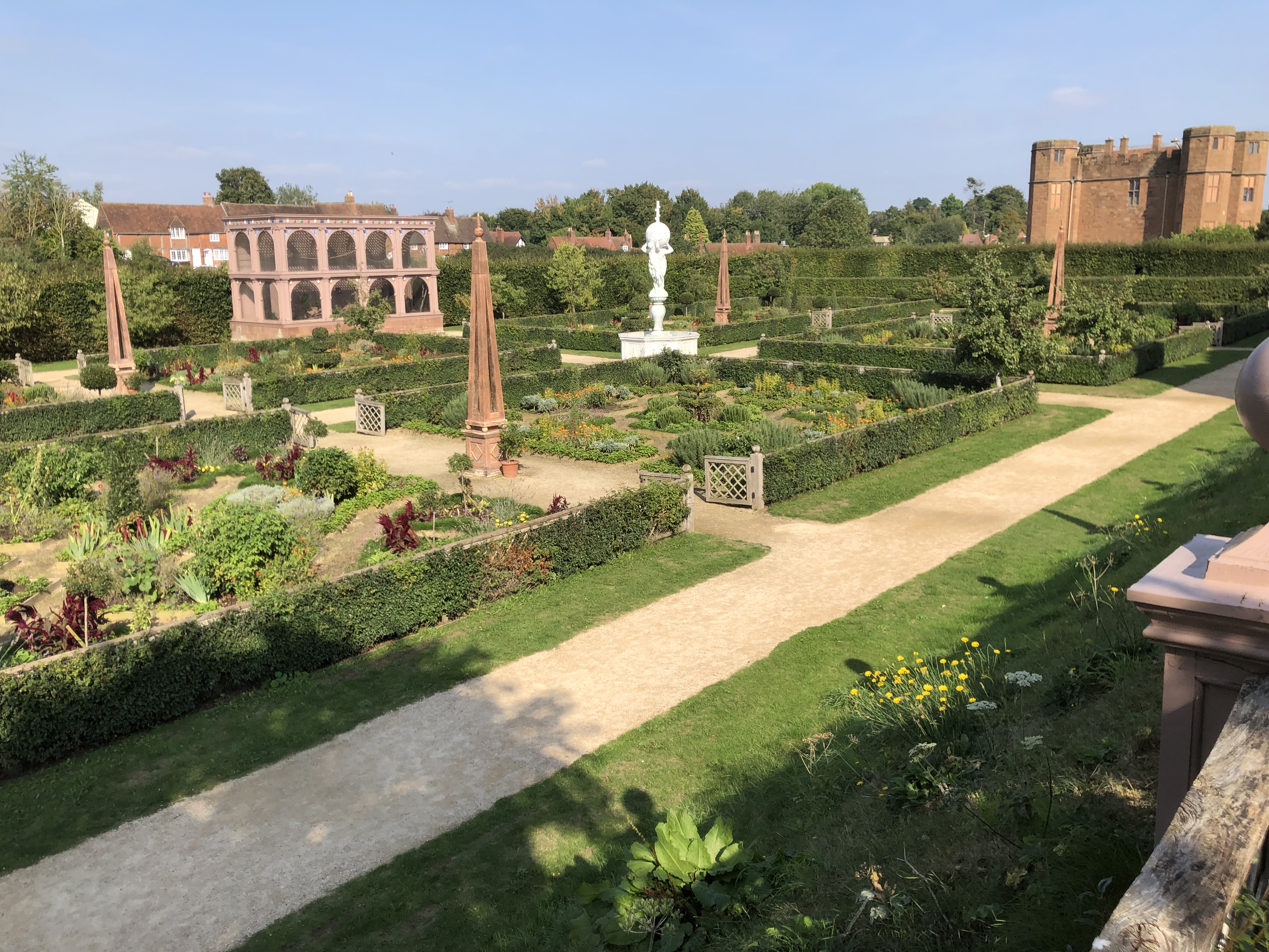 A photograph of a garden with large, square garden beds, paths in between them, and a white central fountain.