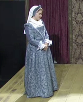 A photo of Julian DeZotti as Cordella. Cordella is a white woman wearing a blue and white outfit and standing with her hands folded in front of herself