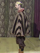 A photo of Jason Gray as Cornwall. Cornwall is a white man with a black goatee. He is wearing a brown and black striped cape and a coronet. He has a sword at his hip.