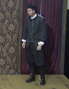 A photo of David Kynaston as Cornwallʼs servant. He is wearing plain dark clothing and standing slightly hunched over.