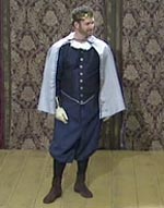 A photo of Paul Hopkins as Gallia. Gallia is wearing a dark blue outfit with a silver cape and golden crown. He is standing with his head turned to the side.