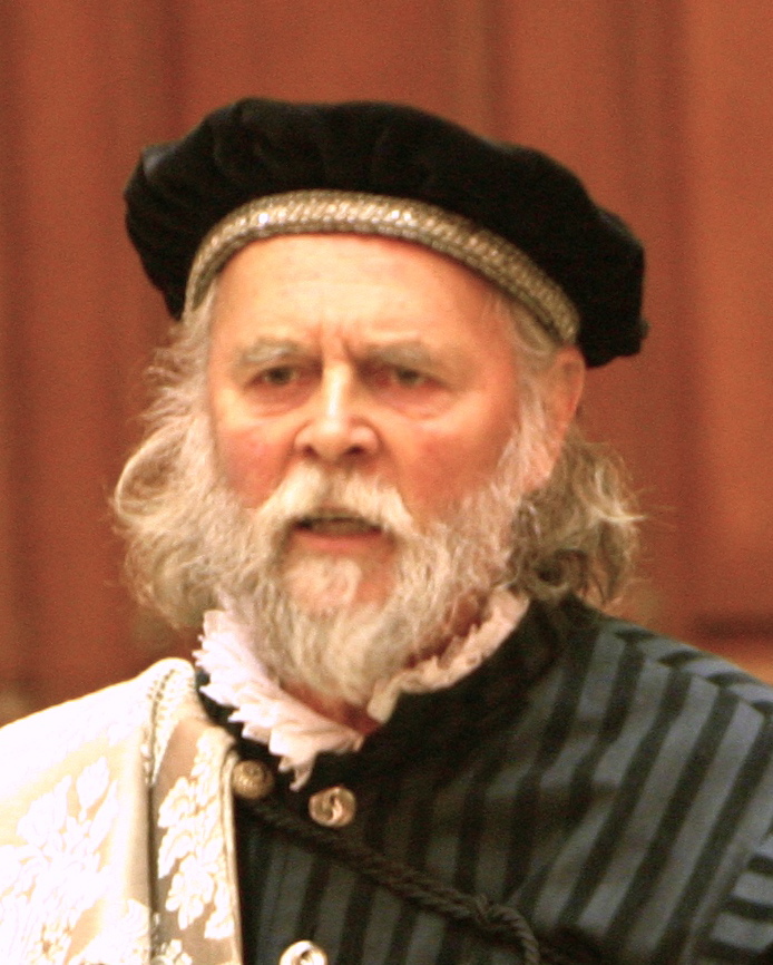 A photo of Peter Higginson as Perillus. Perillus is a white man with shoulder-length grey hair and a beard. He has a furrowed brow and is speaking.